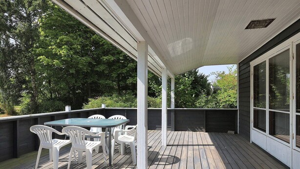 Une terrasse extérieure couverte, dotée d’une table et de chaises, entourée d’une terrasse en bois et d’un mur sombre.