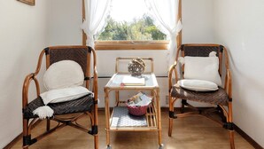 Une pièce comprenant deux fauteuils en osier, une petite table avec un panier, et une fenêtre ornée de rideaux légers.