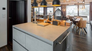 A modern kitchen with a central island, wooden ceiling beams, pendant lighting, and a view of the outdoors through large windows. - Welcoming Villa With Amenities (Trutnov)