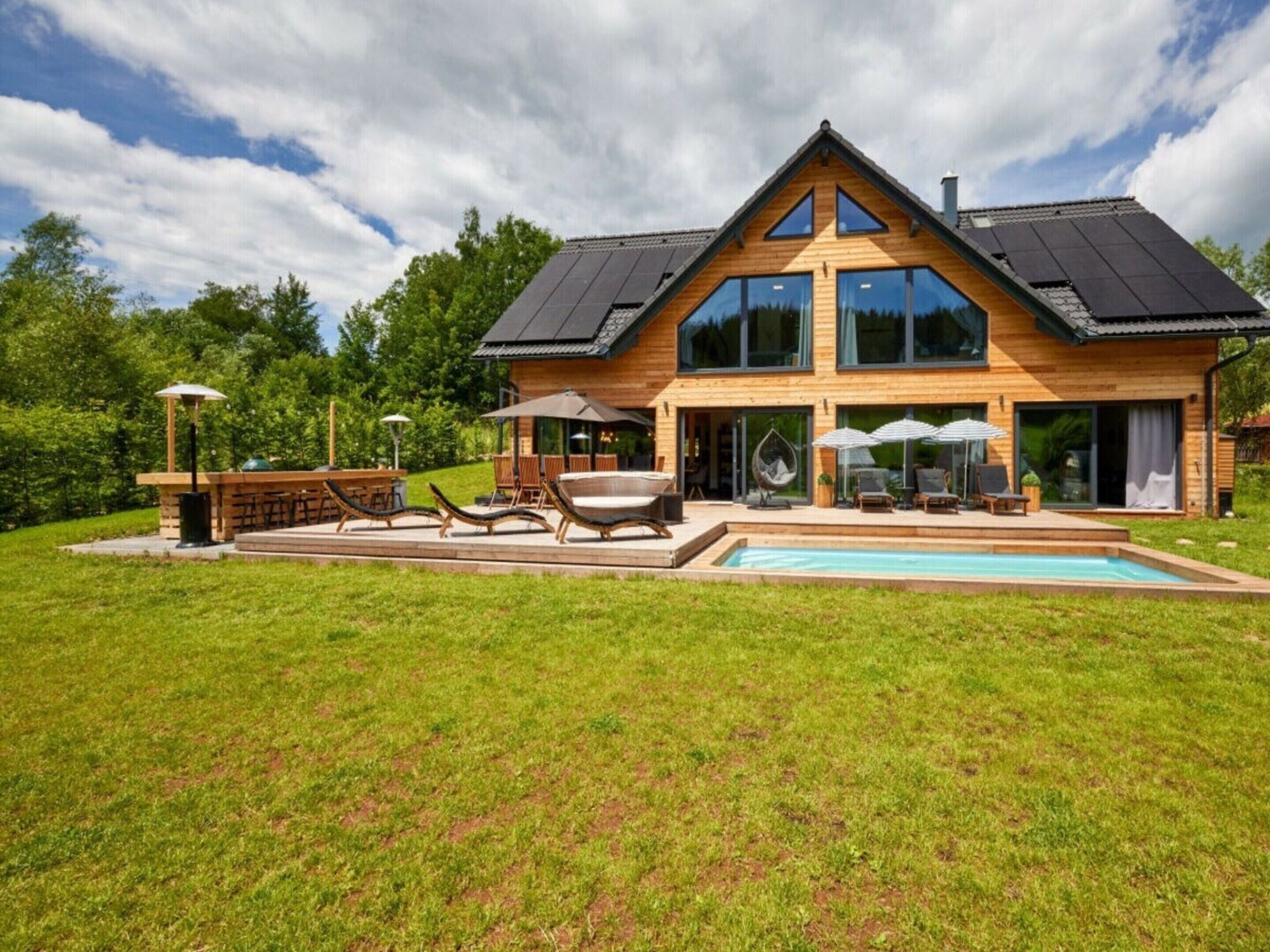 A modern two-story house with a large deck, outdoor seating, and a swimming pool.