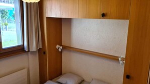 Una camera piccola con un letto singolo, una testiera in legno, una finestra con tende e un termosifone.