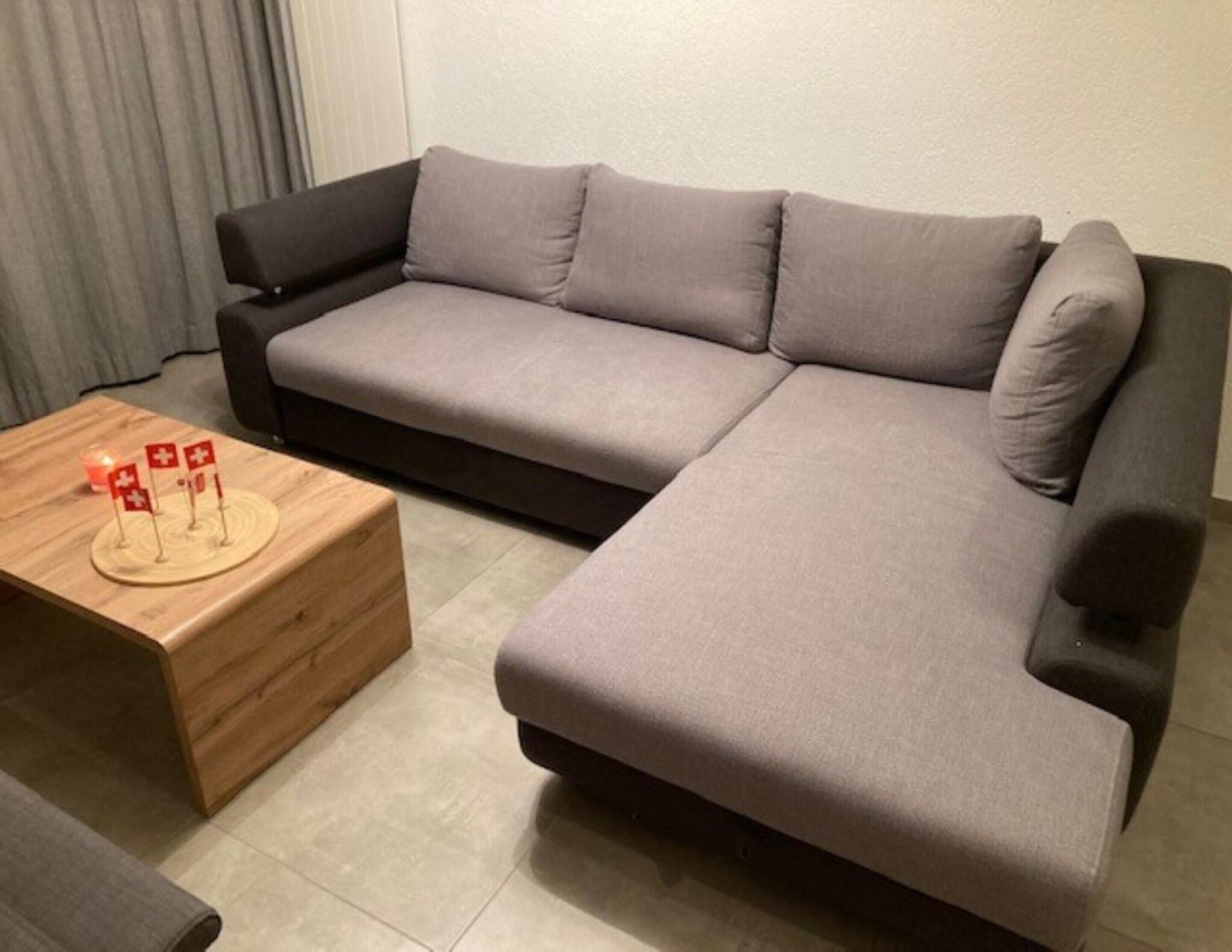 Un salon moderne avec un canapé d’angle gris, une table basse en bois ornée d’un petit drapeau suisse et un mur blanc uni.