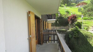 Un balcón con puerta de madera, pared de piedra y vista a un paisaje verde con una casa al fondo.