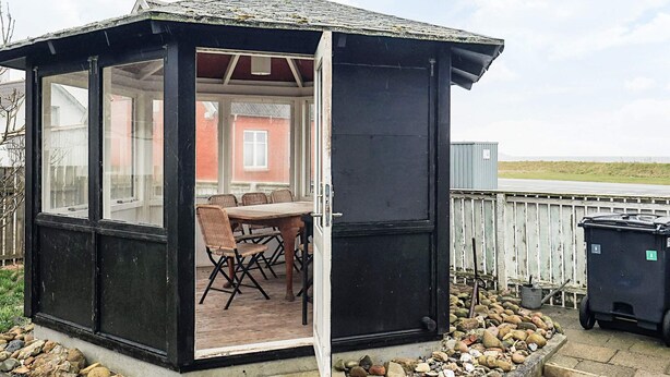 A small, enclosed outdoor area with a table and chairs, a wooden deck, and a view of a red building in the background. - 16 Person Holiday Home in Vestervig (Vestervig)