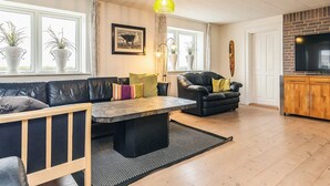 A modern living room with a black leather sofa, a wooden coffee table, and a black armchair. There are potted plants on the windowsill, a framed artwork on the wall, and a television mounted on a brick wall. - 16 Person Holiday Home in Vestervig (Vestervig)