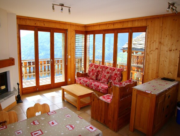 A cozy wooden cabin with a sofa, table, and chairs. Large windows offer a view of the mountains.