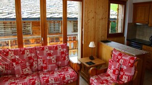 A cozy room with a wooden sofa and armchair, a small table with a lamp, and a kitchen area with a stove and cabinets. - Cozy Apartment With Rhone Valley View (Nendaz)