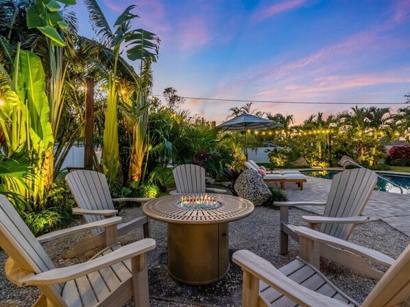 A patio with Adirondack chairs, a round table with a fire feature, and a pool in the background.