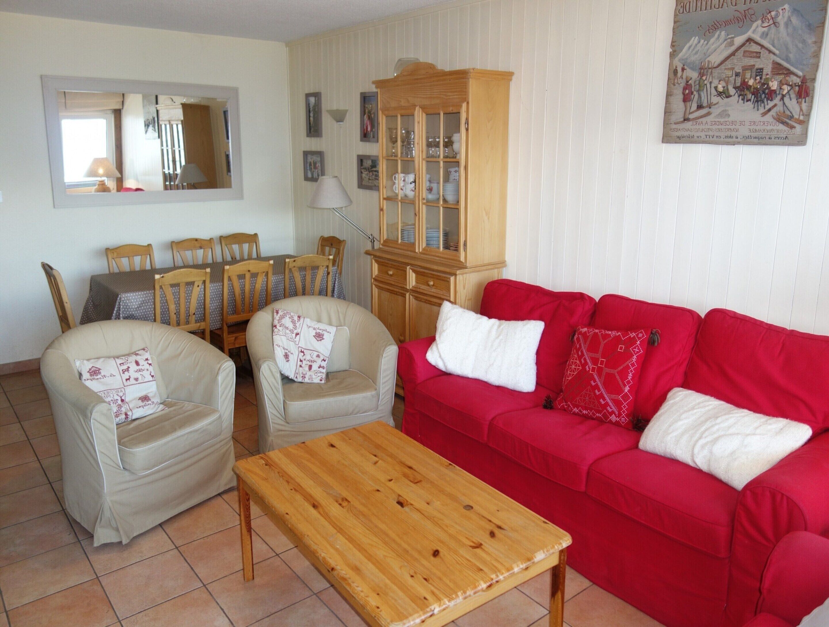 Ein Wohnzimmer mit einem roten Sofa, zwei Sesseln, einem Holztisch, einer Holzkommode und einem Esstisch mit Stühlen.