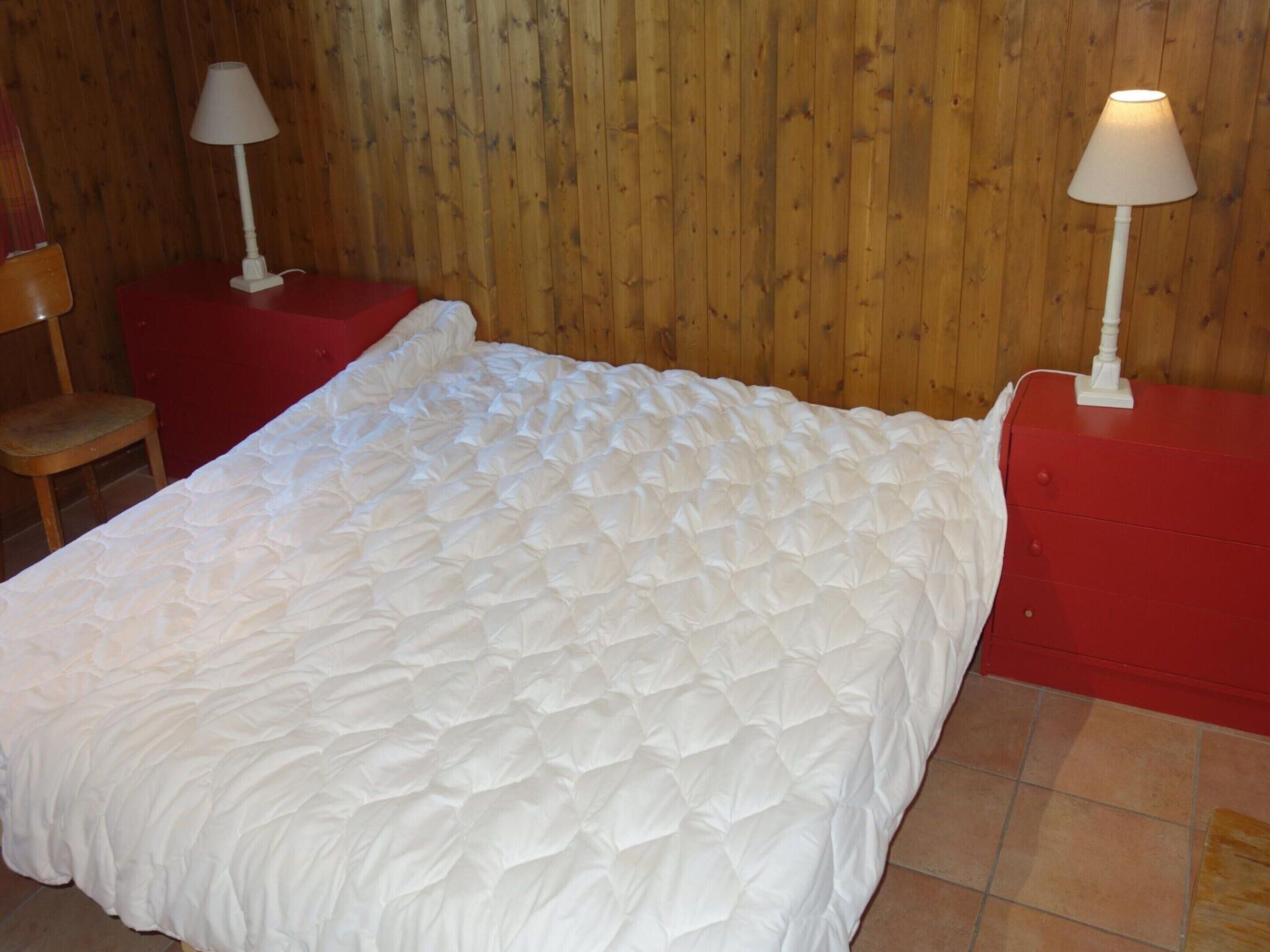 A neatly made bed with a white quilt, a wooden wall, and a red nightstand with a lamp.