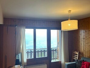 A room with wooden walls, a large window with curtains, a hanging lamp, a chair, and a view of the outdoors. - Studio With a Hilltop View (Veysonnaz)