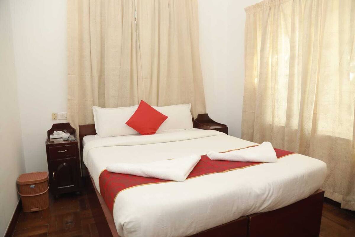 A neatly made bed with a red and white bedspread, a wooden nightstand with a lamp, and a small trash bin.