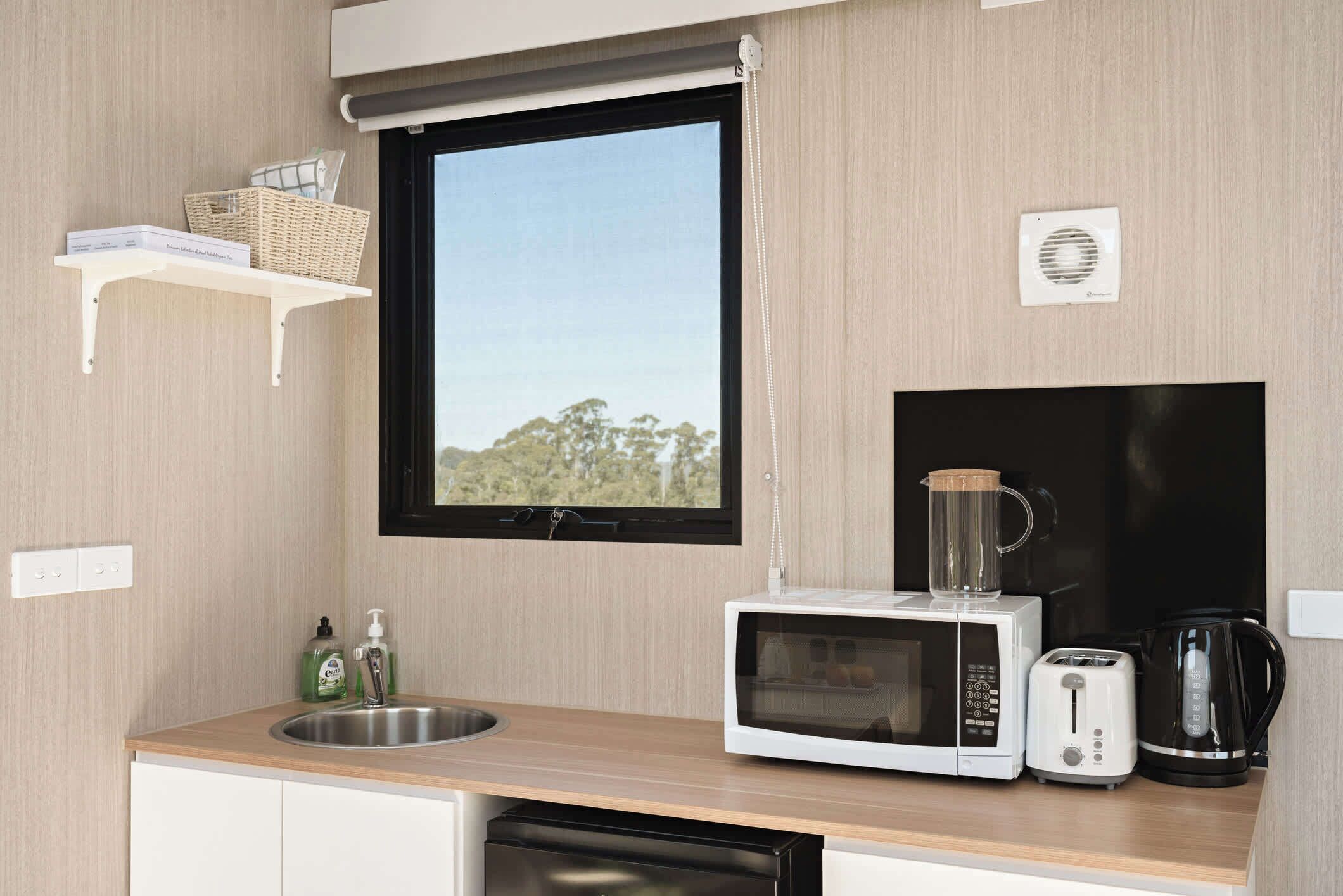A compact kitchen with a microwave, toaster, and coffee maker on a wooden countertop, a window with a view, and a small shelf with a basket.