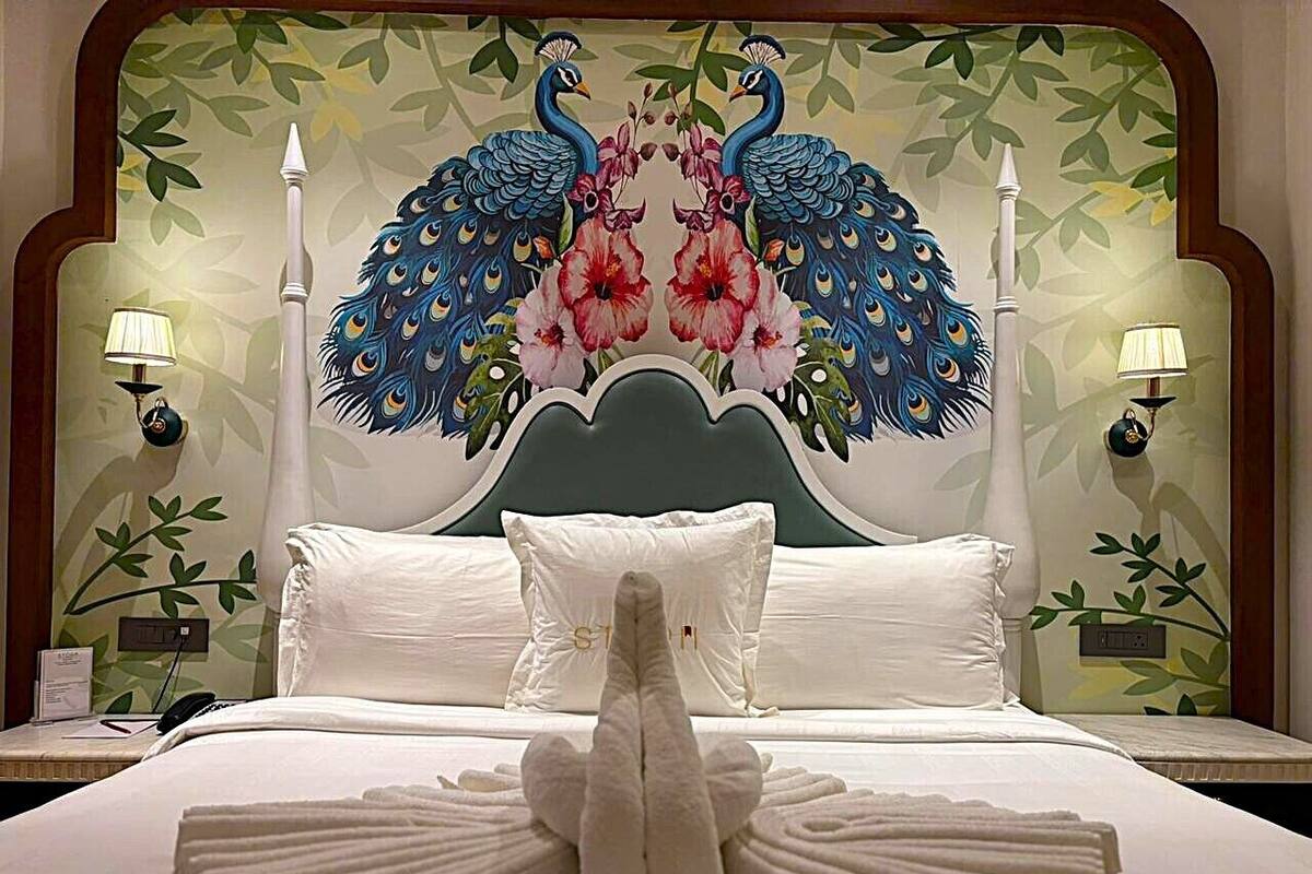 Ein Hotelzimmer mit einem großen Bett, weißer Bettwäsche und einem Wandbild eines Pfauens auf dem Kopfteil. Zwei Nachttischlampen und ein kleiner Tisch mit einer Lampe sind ebenfalls vorhanden.