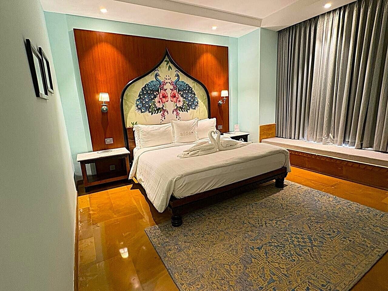 A bedroom with a large bed, a decorative headboard, bedside lamps, a window with curtains, and a patterned rug.