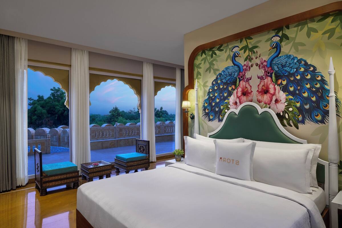 Ein Hotelzimmer mit einem großen Bett, Blick auf einen Garten durch Rundbogenfenster und einem Wandgemälde mit Pfauen und Blumen.