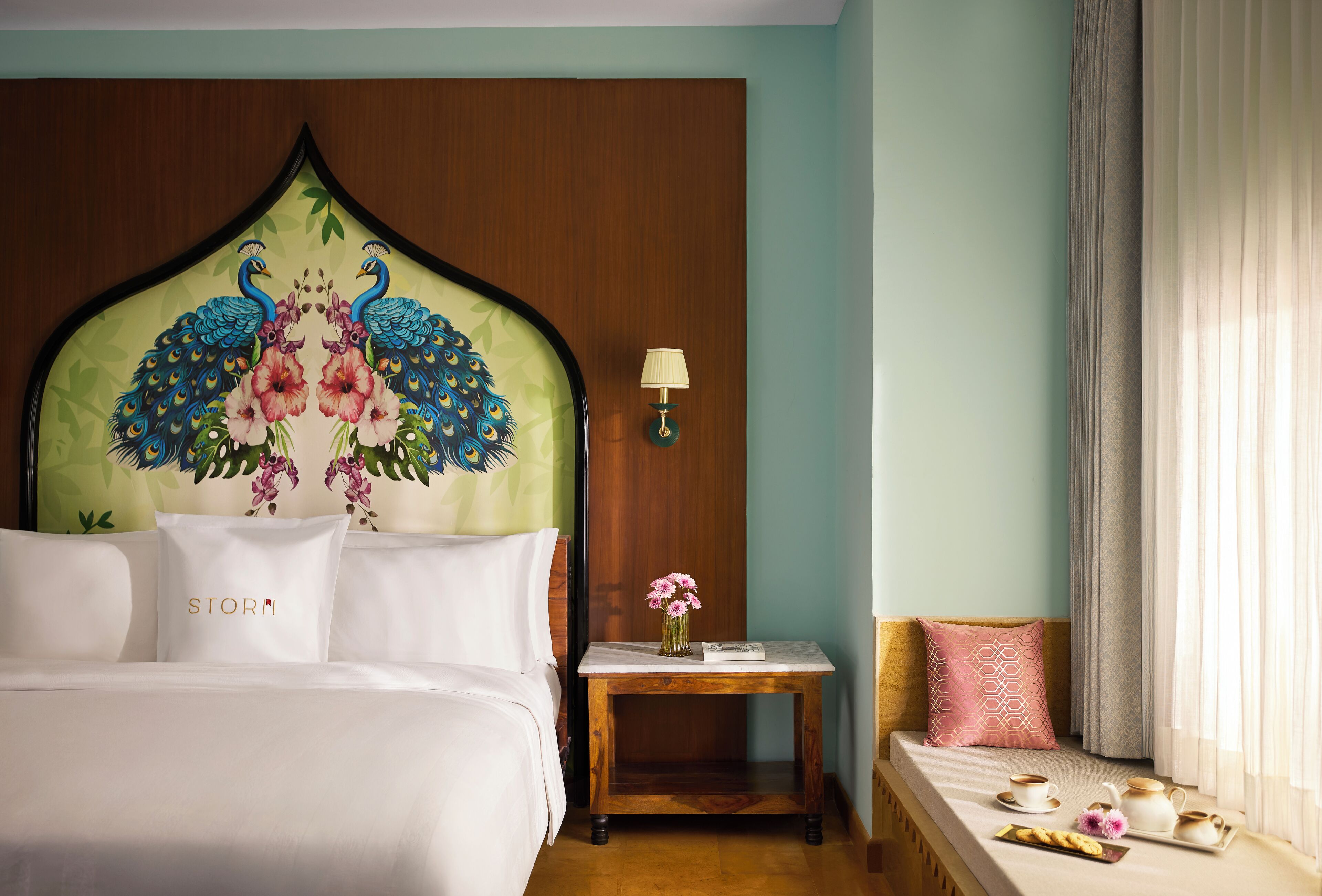 Una habitación de hotel con una cama grande, una mesita de noche con lámpara, una pequeña mesa con un jarrón de flores y una ventana con cortinas.