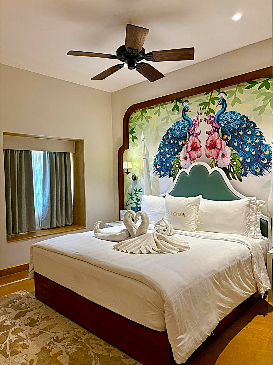 A hotel room with a bed, a ceiling fan, a window with curtains, and a wall with a peacock mural.
