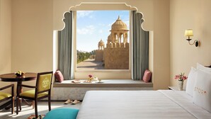 Habitación de hotel con un ventanal amplio que ofrece vistas a un edificio histórico, una cama, un escritorio con silla, y una pequeña mesa con un florero.