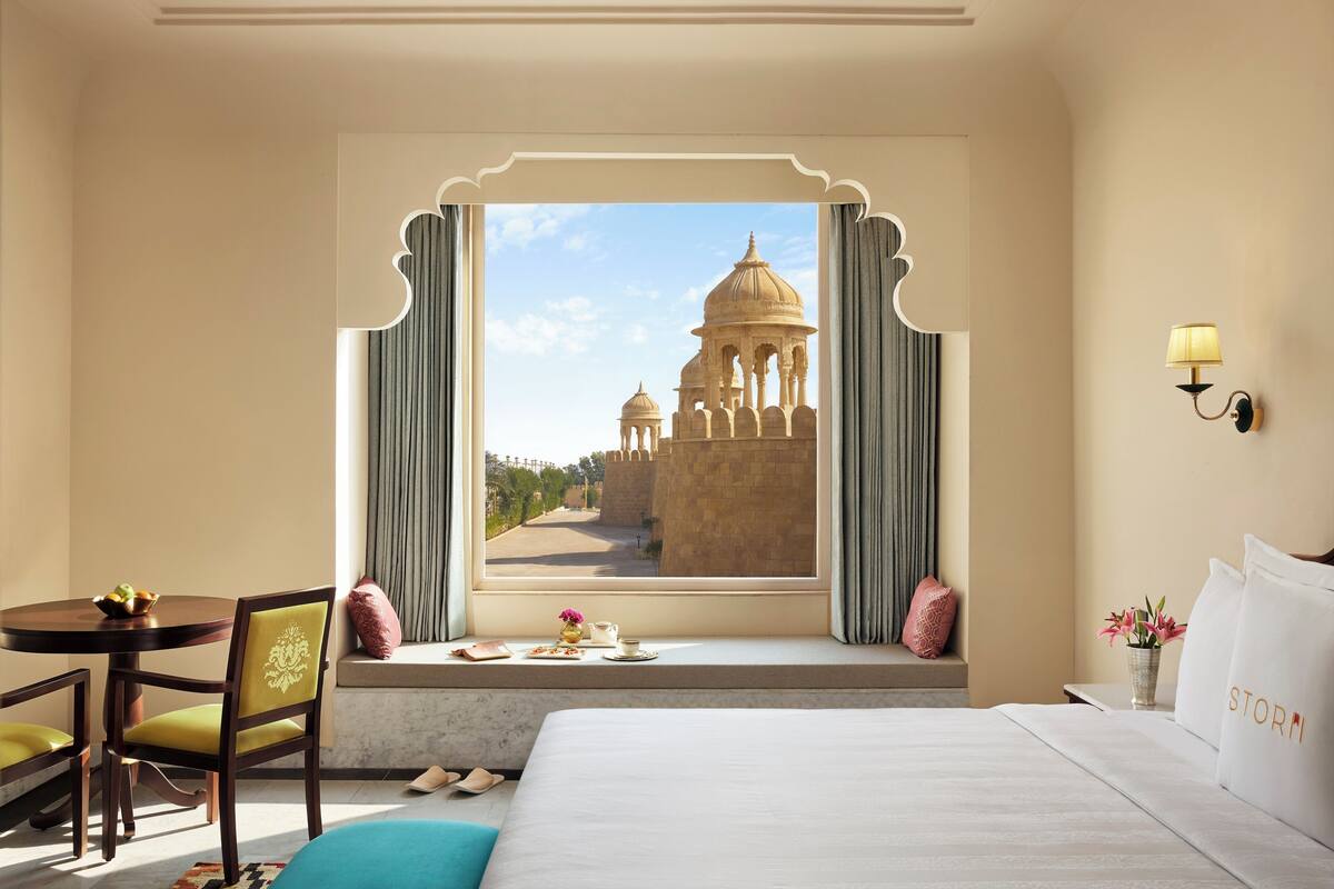 Ein Hotelzimmer mit großem Fenster und Blick auf ein historisches Gebäude, ein Bett, ein Schreibtisch mit Stuhl und ein kleiner Tisch mit Blumen.
