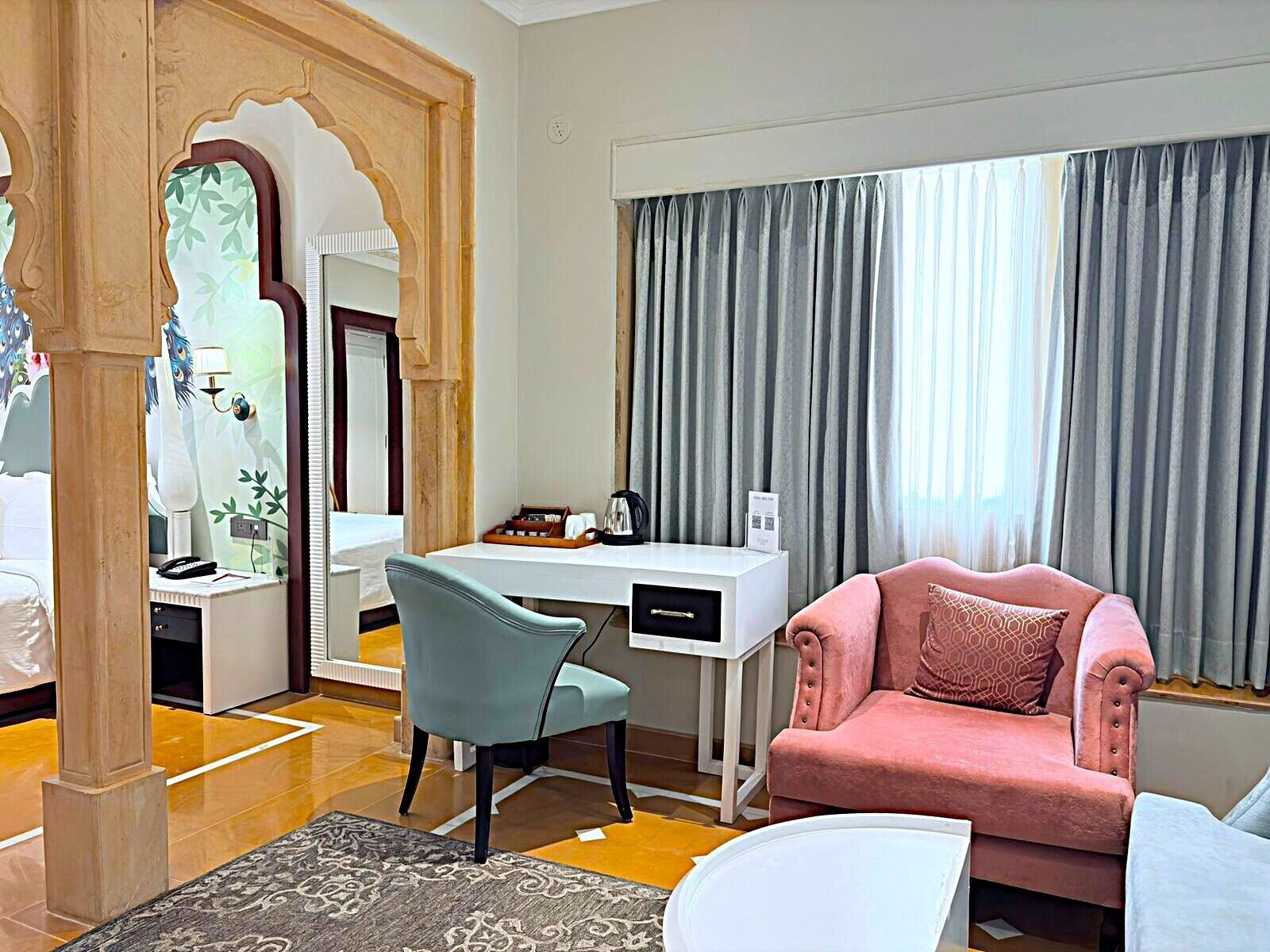 Une chambre d’hôtel avec un lit, un bureau et une chaise, un fauteuil rose, une grande fenêtre avec des rideaux et un miroir décoratif.