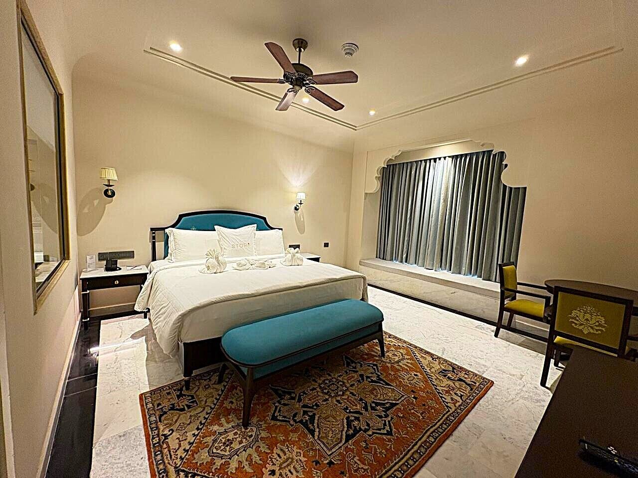 A hotel room with a bed, a bench, a desk, and a ceiling fan.