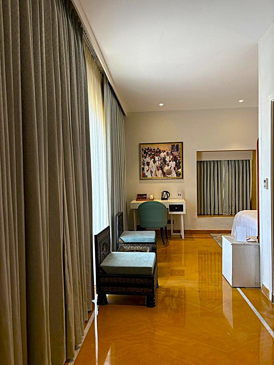 A hotel room with a wooden floor, a desk with a chair, a bed, and a large window with curtains.