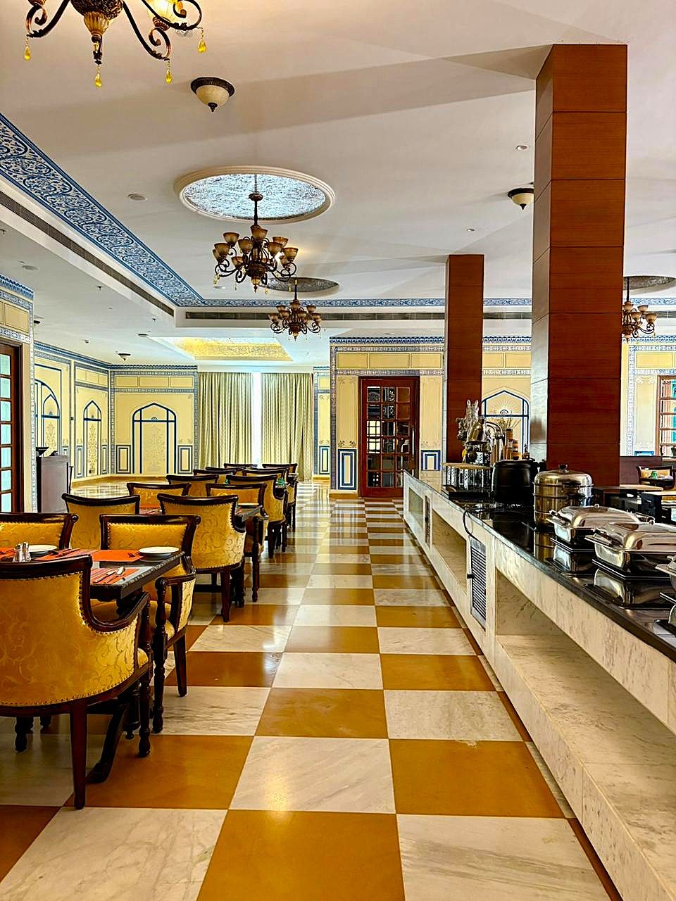 Un hall de hotel con piso a cuadros, comedor con sillas amarillas, un mostrador de buffet y candelabros decorativos.