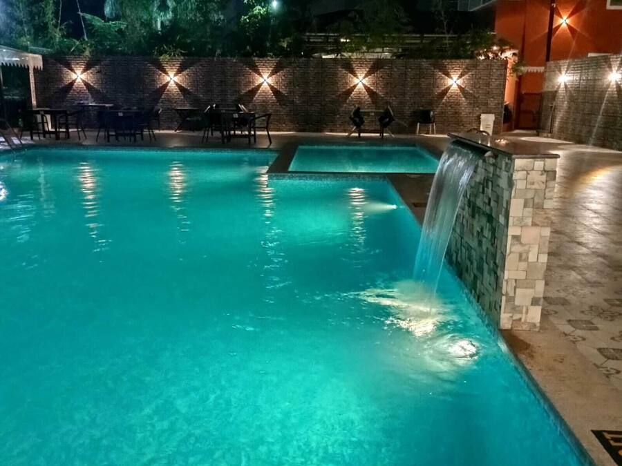 Una piscina con cascada, rodeada de mobiliario exterior y un muro de ladrillo con luces iluminadas.