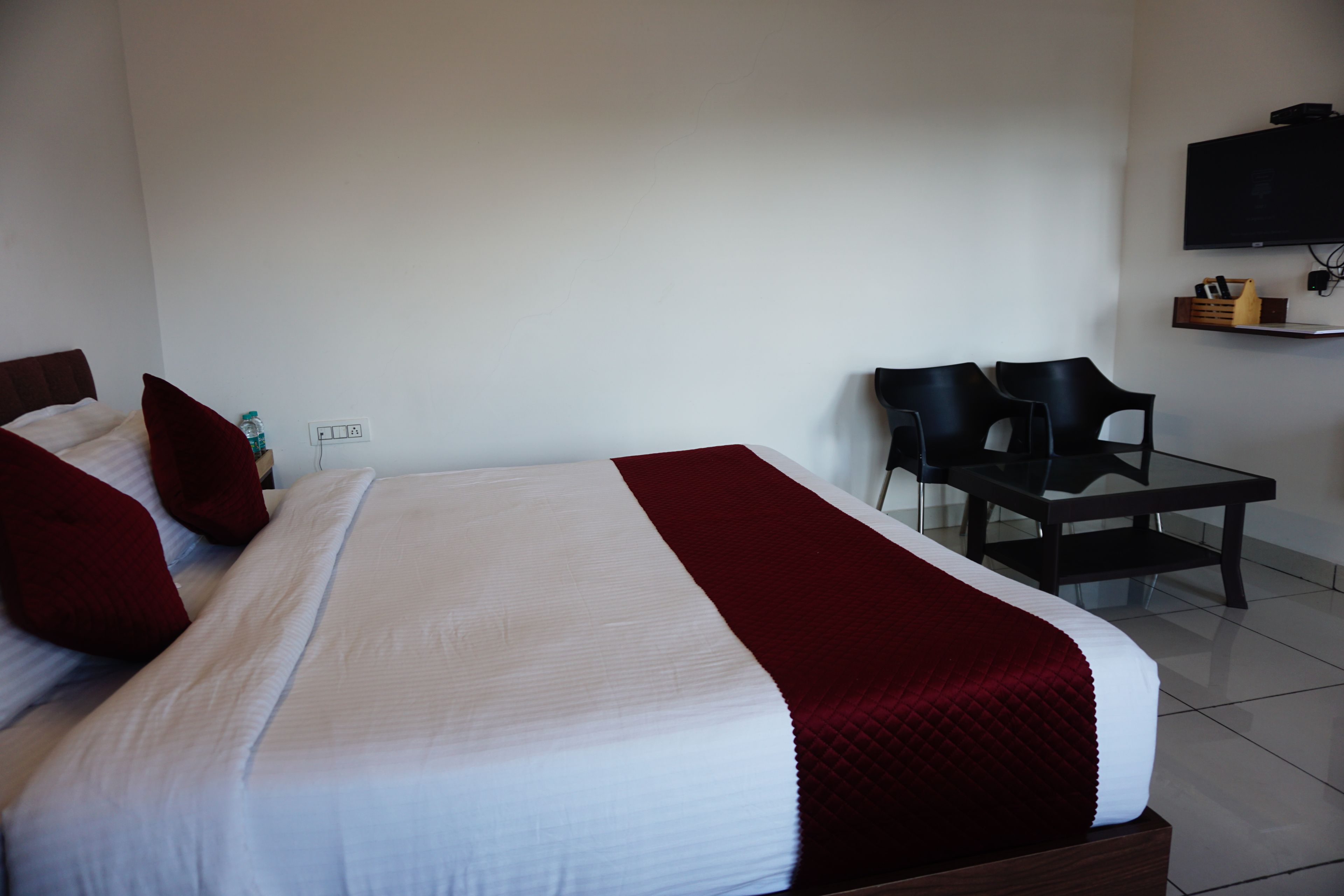 Quarto de hotel com cama, escrivaninha, duas cadeiras, televisão e uma tomada na parede.