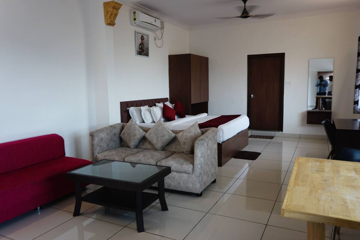 A modern hotel room with a bed, a red sofa, a grey sofa, a glass coffee table, and a wooden table.