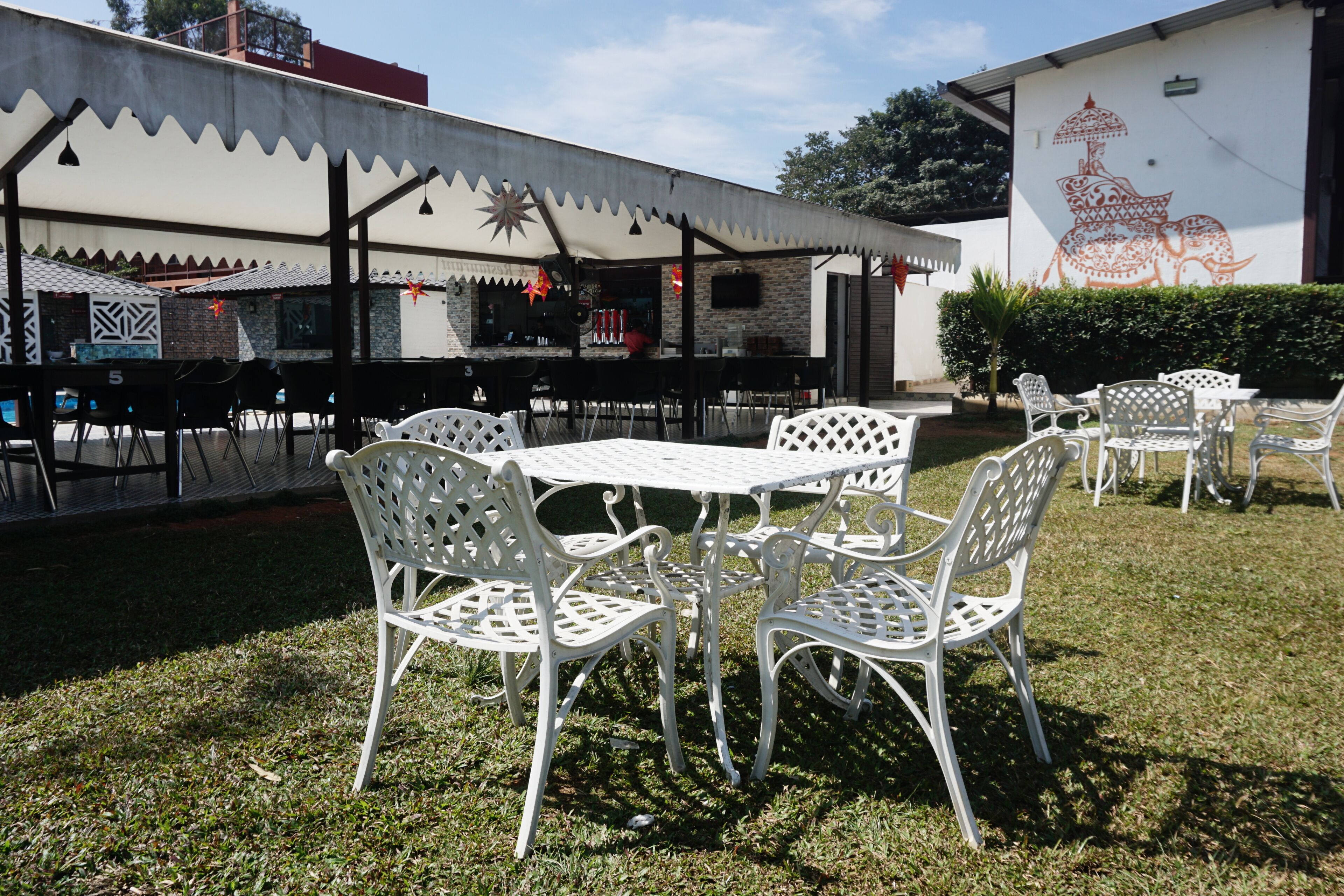 An outdoor seating area with white metal chairs and tables, a covered patio, and a mural on the wall.