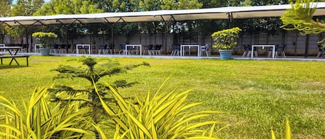 Un espacio al aire libre techado con mesas y sillas, rodeado de césped y plantas.