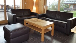 A room with two black leather sofas, a wooden coffee table, and a large window with a view of a deck and trees. - Apartment With Panoramic Views (Veysonnaz)