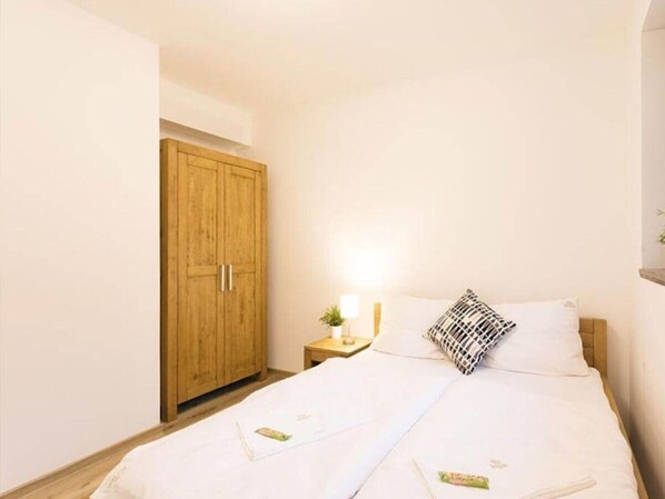 A bedroom with a wooden wardrobe, a bedside table with a lamp, and a bed with white bedding and a patterned pillow. - Comfortable Apartment With Living Room (Harrachov)