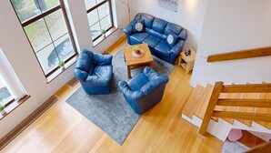 Un vaste espace de vie agrémenté d’un canapé en cuir bleu, de deux fauteuils bleus et d’une table basse en bois. De grandes fenêtres laissent entrer la lumière naturelle, et un escalier en bois est visible sur la droite.