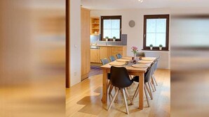 Un espace de salle à manger moderne avec une table et des chaises en bois, une cuisine en arrière-plan et de grandes fenêtres.
