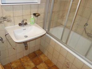 Un baño con lavamanos blanco, cabina de ducha de vidrio y suelo de baldosas marrones.