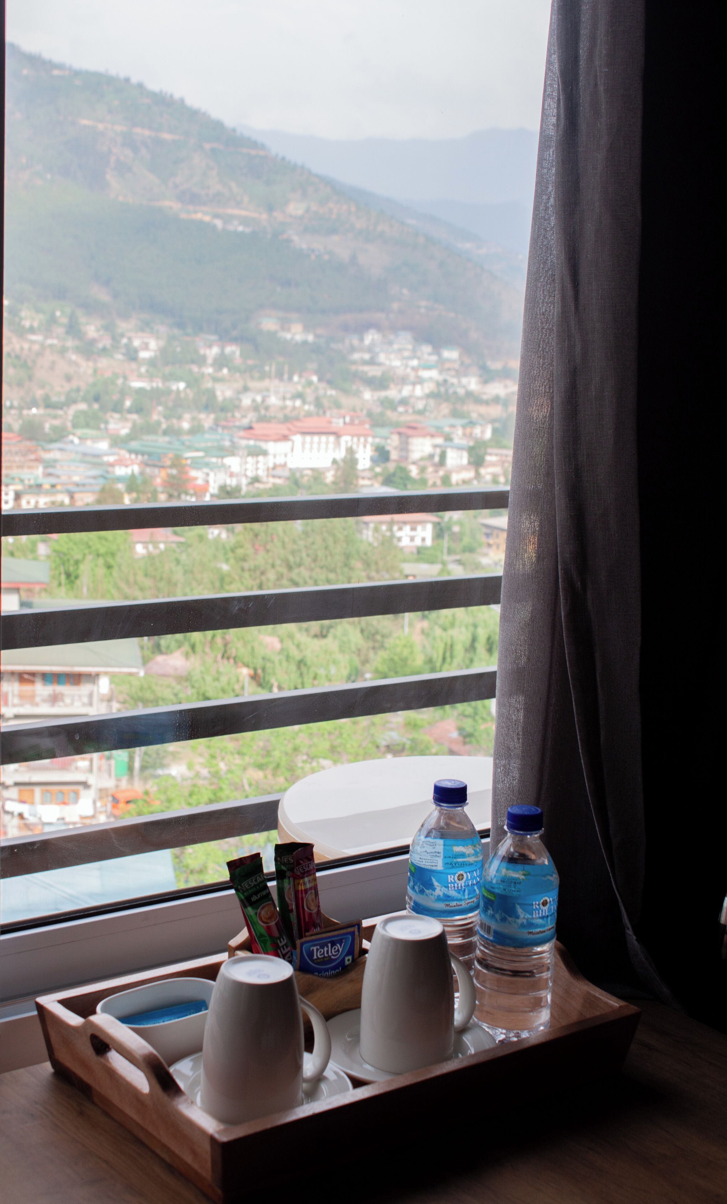 A tray with cups, Tetley tea bags, and bottled water on a table by a window overlooking a mountainous landscape.