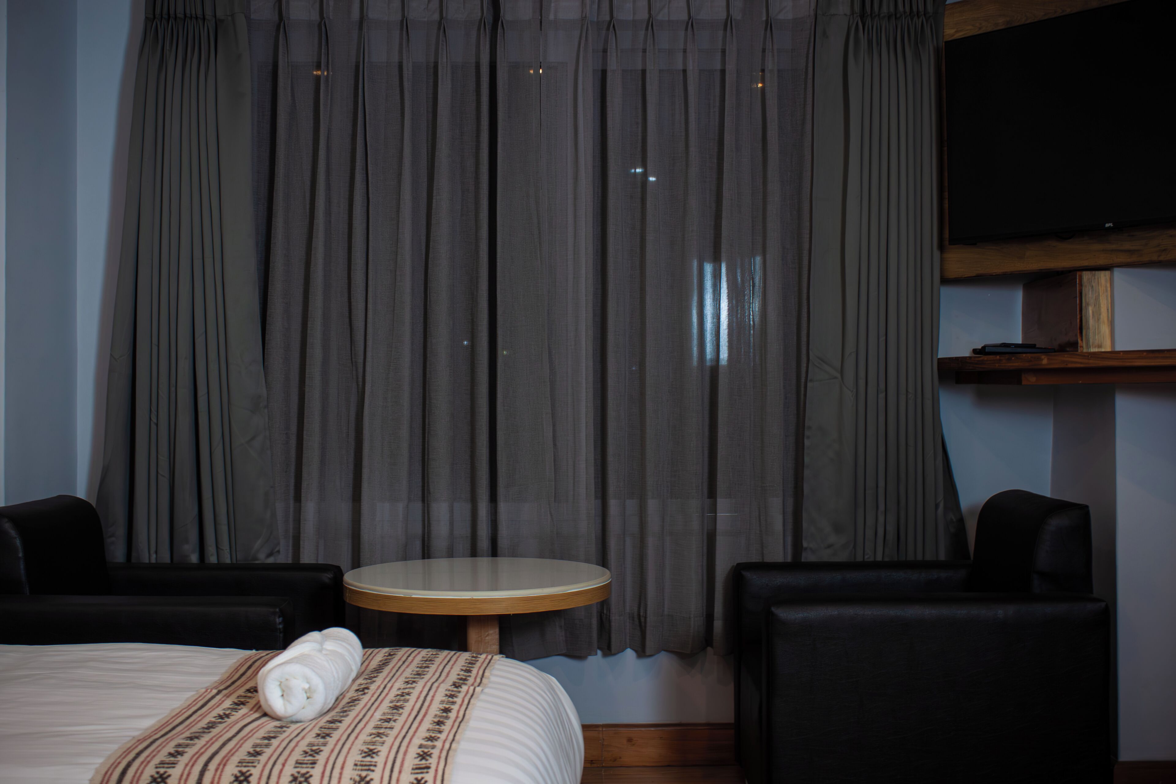 A hotel room with a bed, a round table, two black armchairs, and a TV mounted on the wall.