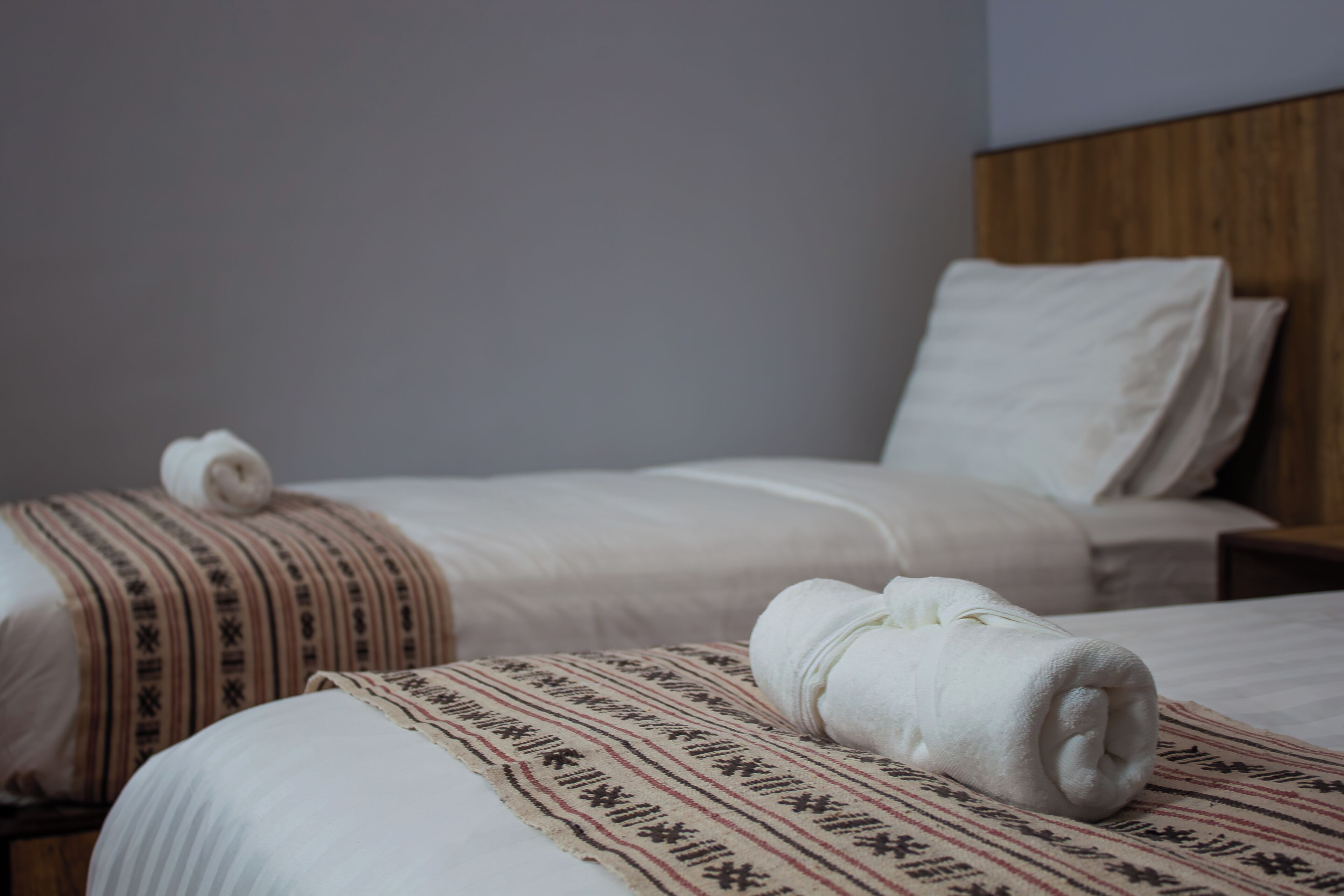 Two beds with white linens and patterned blankets, a wooden headboard, and a plain wall.