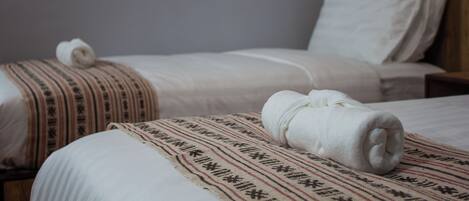 Two beds with white linens and patterned blankets, a wooden headboard, and a plain wall.