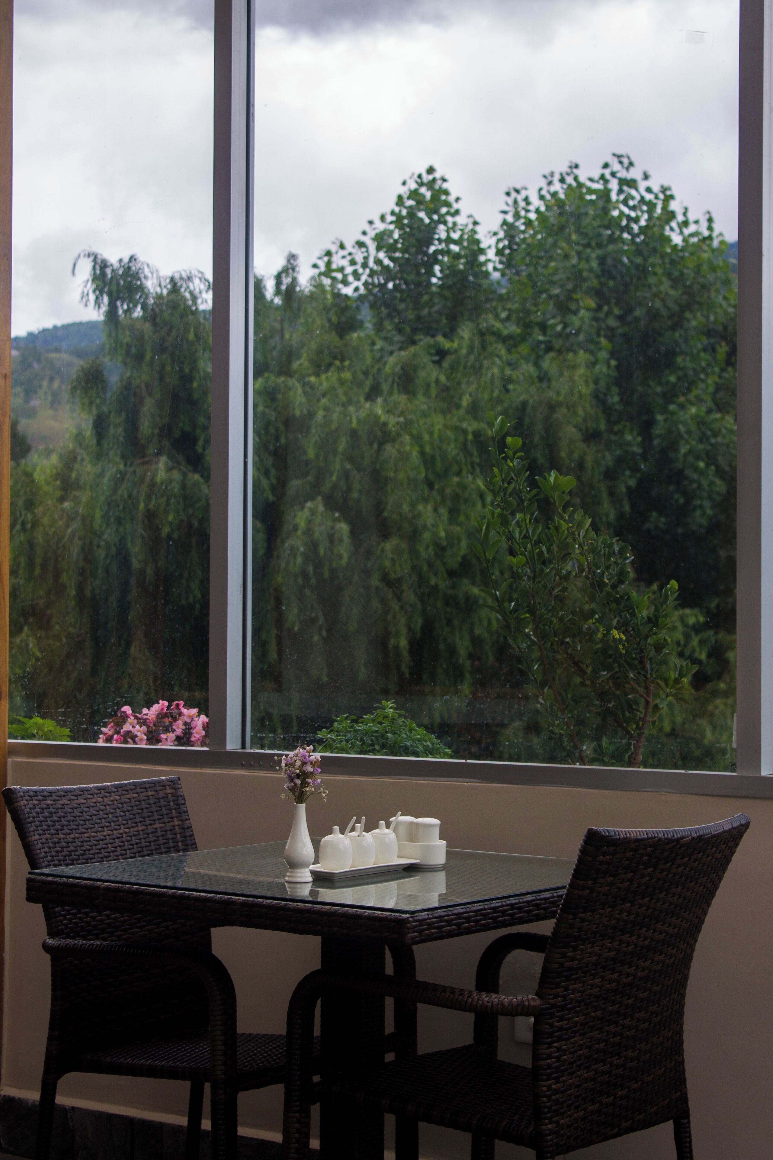 Una mesa de comedor con sillas y un florero, con vista a un gran ventanal que muestra árboles y colinas.