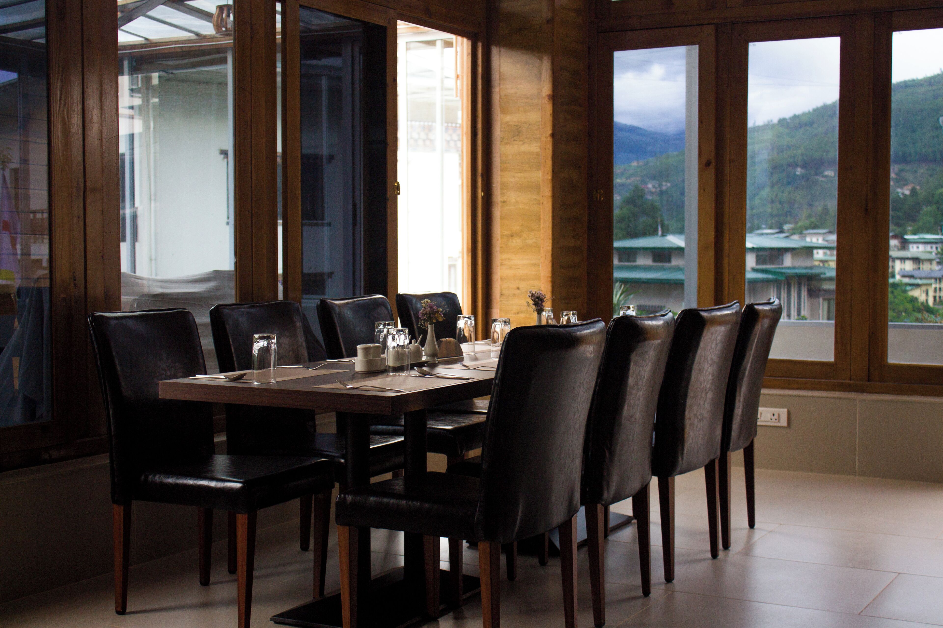 A dining area with a wooden table set for six, surrounded by black chairs, with a view of a mountainous landscape through large windows.