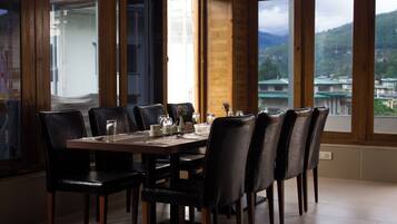 Un coin repas avec une table en bois dressée pour six personnes, entourée de chaises noires, et offrant une vue sur un paysage montagneux grâce à de grandes fenêtres.