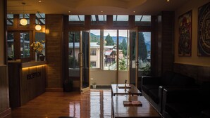 A reception area with a wooden reception desk, a seating area with a table and chairs, and large windows offering a view of buildings and mountains. - Hotel Bhutan Home Thimphu (Thimphu)