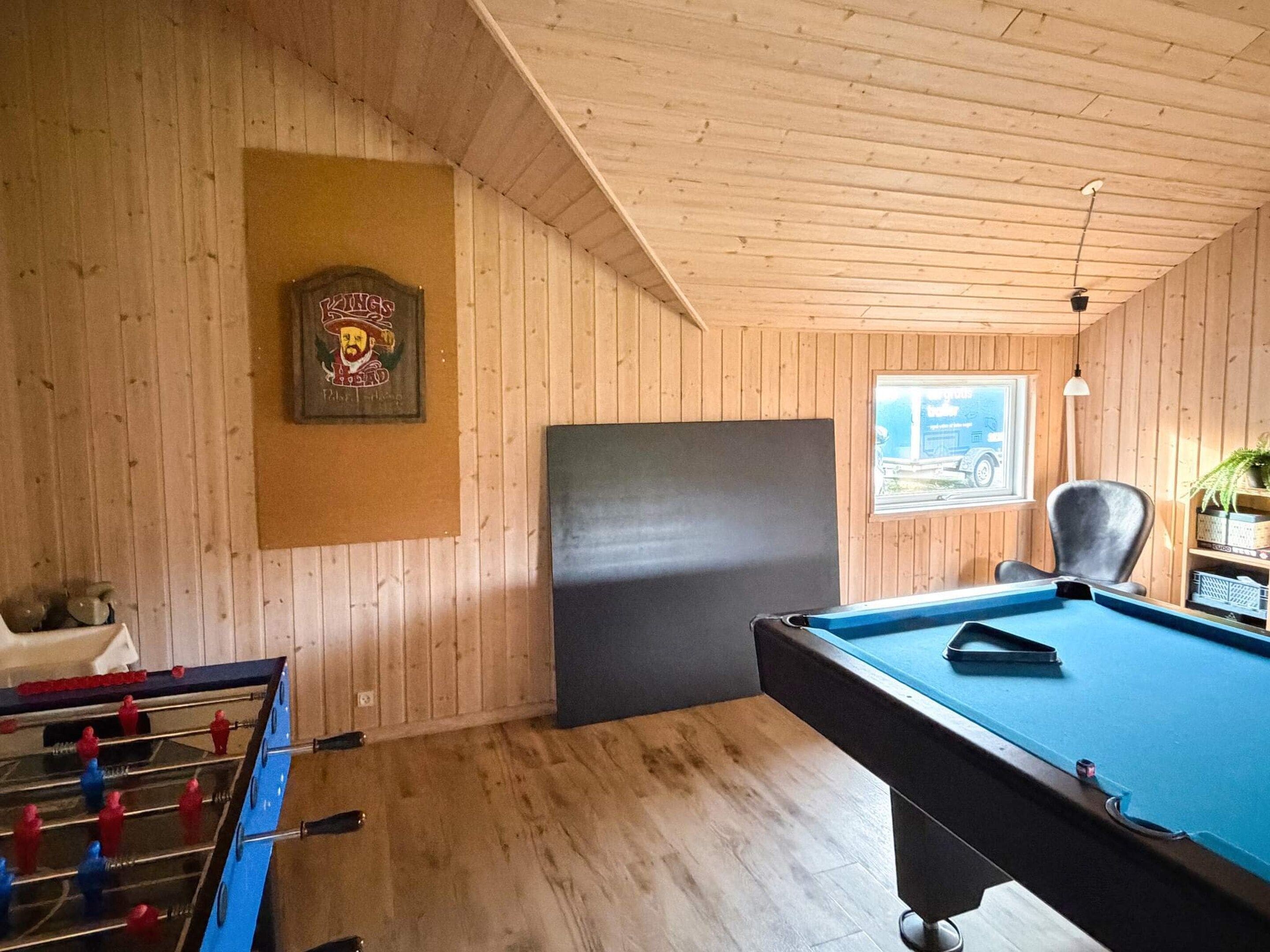 Une pièce avec un baby-foot, une table de billard, des murs en bois, un tableau encadré et une fenêtre donnant sur un beau paysage.