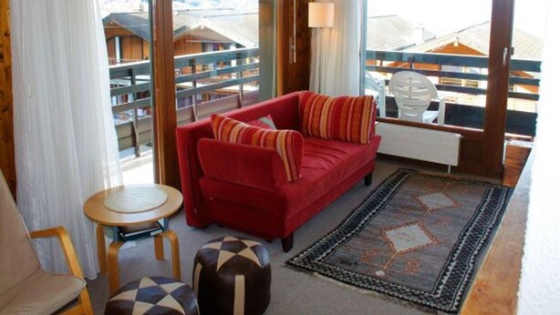 Une pièce chaleureuse avec un canapé rouge, un pouf à motifs, une petite table ronde et une vue sur une terrasse en bois et une bibliothèque, le tout visible à travers de grandes fenêtres.