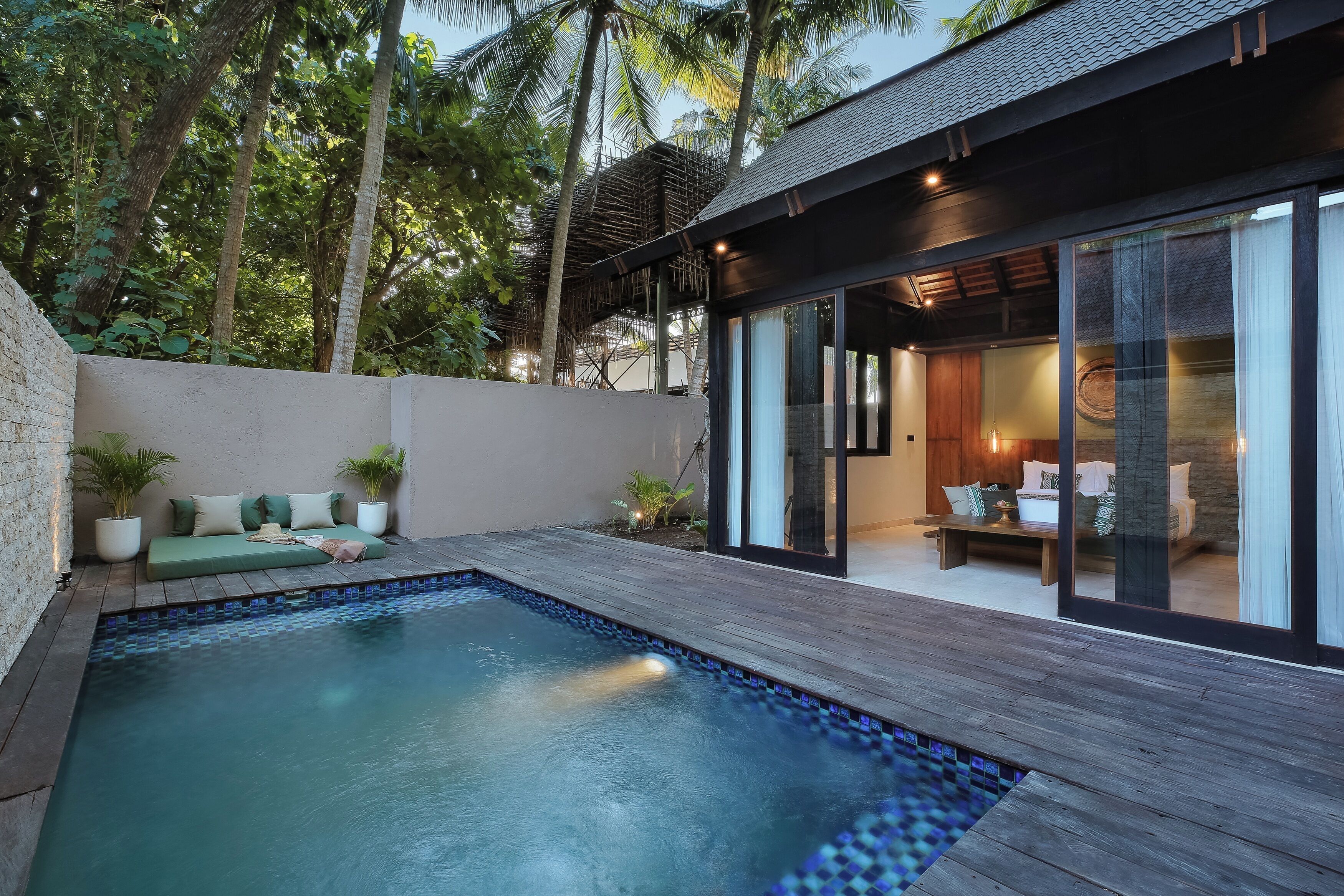 A modern outdoor pool area with a wooden deck, a seating area with cushions, and a view of the interior through large glass doors.