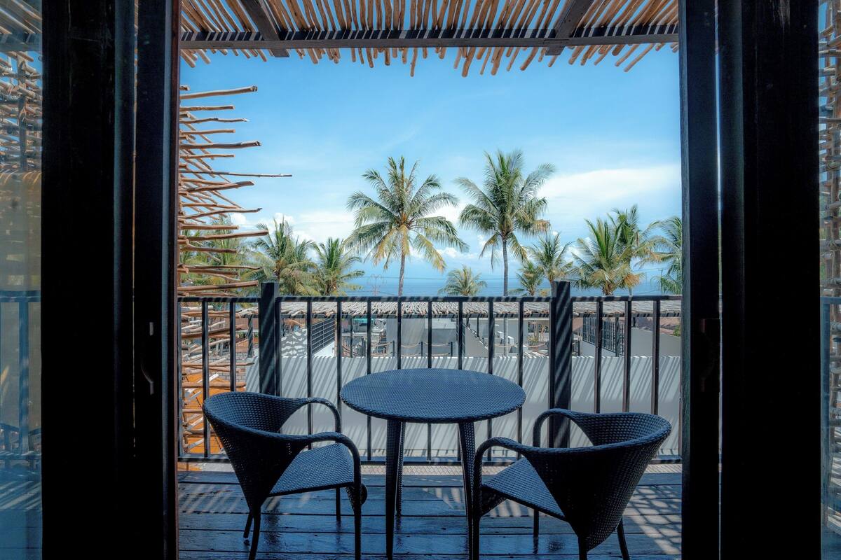 A balcony with a round table and two chairs, overlooking palm trees and a clear sky.